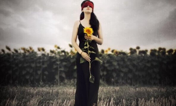 Een geblinddoekte vrouw die voor een veld met zonnebloemen staat en zelf ook een zonnebloem vasthoudt